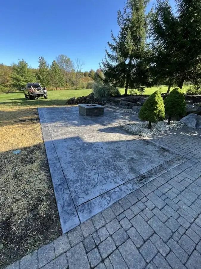 Stone patio with a circular fire pit in a grassy backyard, bordered by shrubs and trees