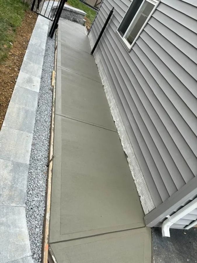 Concrete walkway beside a gray-sided house, bordered by gravel and a narrow stone edge.