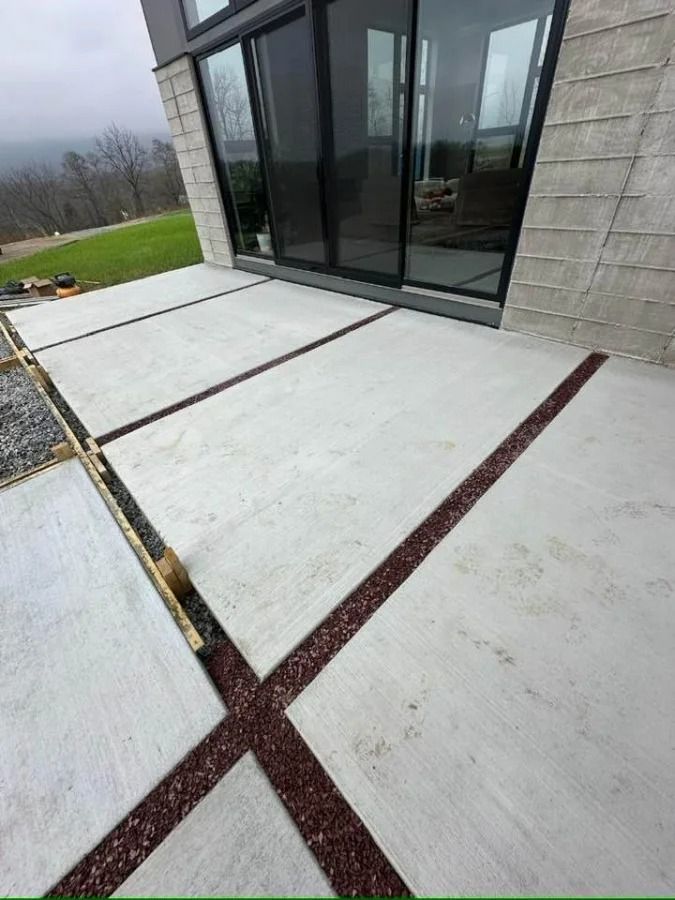 Concrete patio with dark expansion joints beside a modern glass sliding door and gray stone wall.