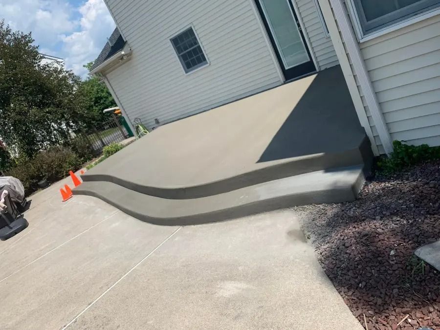 Concrete driveway ramp with curved steps beside a white house; orange cones mark the edge.