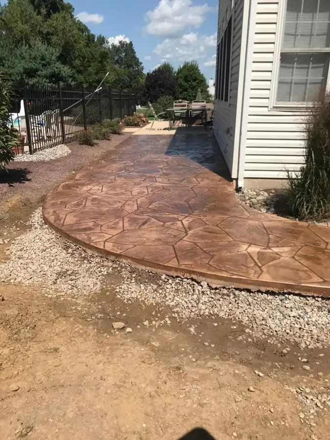 Stamped concrete walkway beside a white house, with dirt edges and a fenced yard under a sunny sky