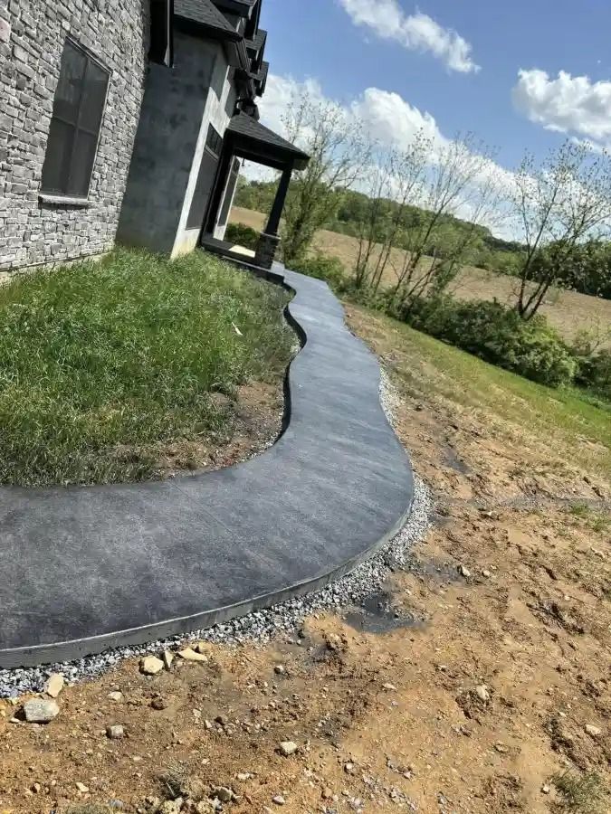 Curving concrete walkway beside a stone house, with dirt and grass along the edges.