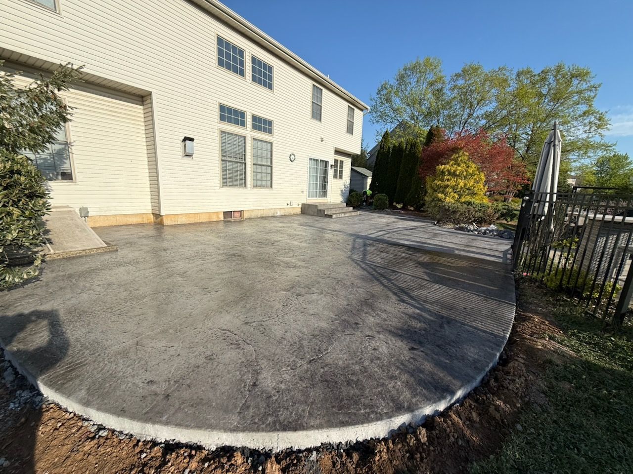 Curved concrete patio beside a beige house, bordered by shrubs and a black fence under a clear sky