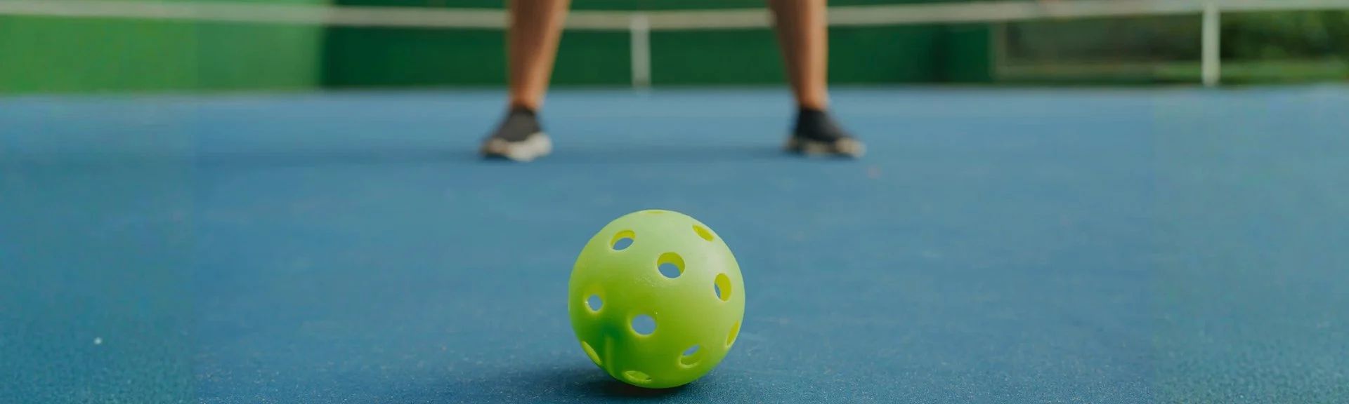 Green pickleball ball on a blue court with a player’s legs blurred in the background