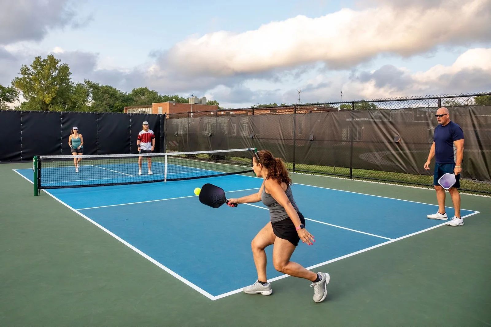 Two women playing pickleball on a blue outdoor court, with two others watching nearby.