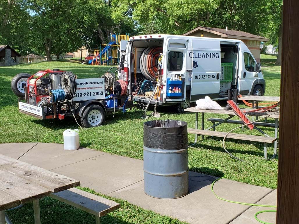 A cleaning service van parked at a park with equipment set up; a trash can and picnic table in the foreground.