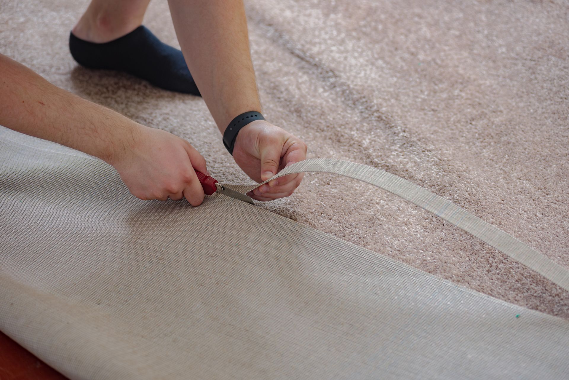 Person using a utility knife to cut a strip from a roll of beige carpet.