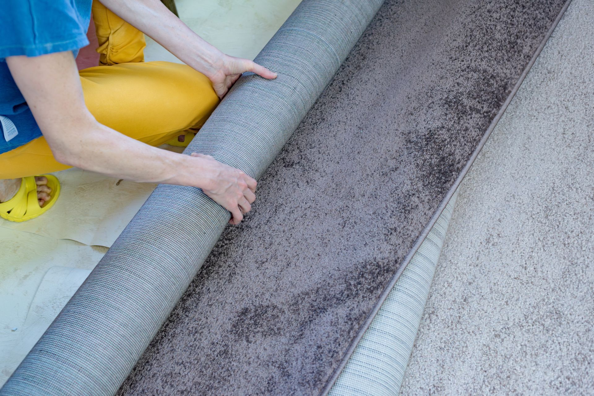 Person rolling up a gray rug, revealing a lighter carpet underneath.