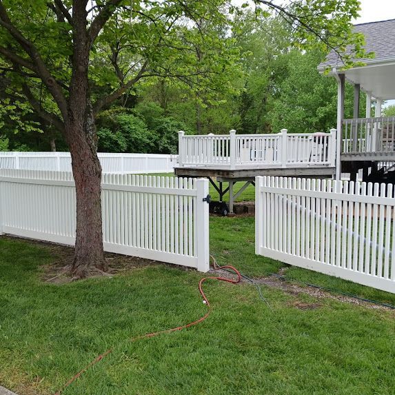 White picket fence around a grassy yard with a tree and raised deck, gate open in front