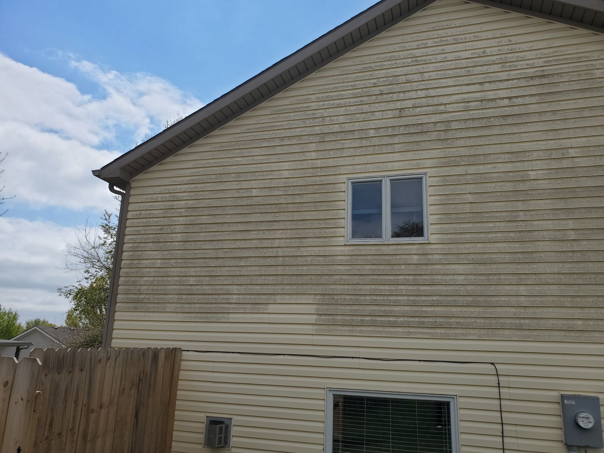 Dirty siding on a two-story beige house with a window, against a partly cloudy sky.