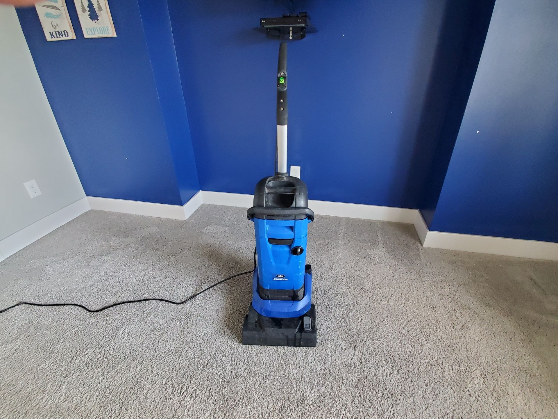 Blue vacuum cleaner standing on gray carpet in front of a blue wall.