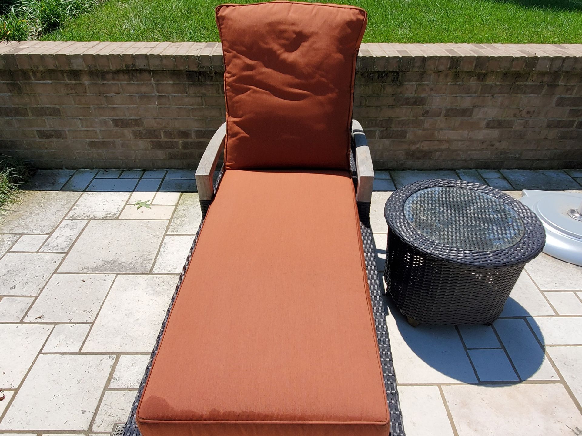 A chaise lounge with rust-colored cushions and a small dark wicker side table on a patio.