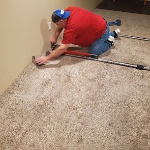 Person kneels, installing carpet along a wall, using a knee kicker and tools.