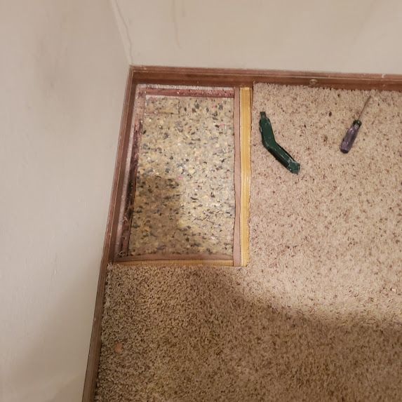 Carpet removal in progress; exposed padding, wood trim, screwdriver, and utility knife on floor.