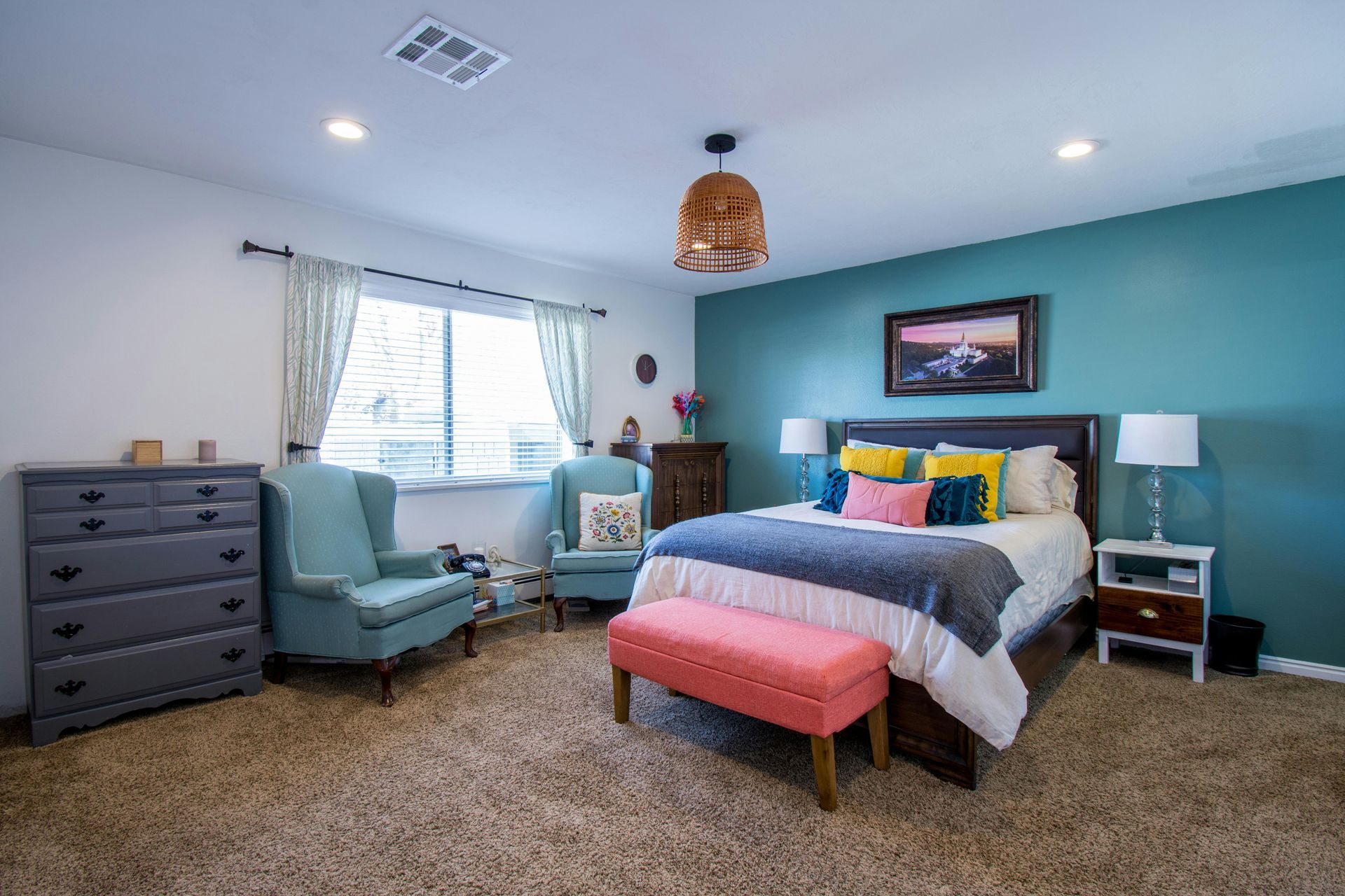 Bedroom with teal accent wall, bed, chairs, dresser, window with curtains, and rug.