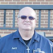 Man in blue shirt and sunglasses stands in front of a brick wall, wearing a cross necklace.