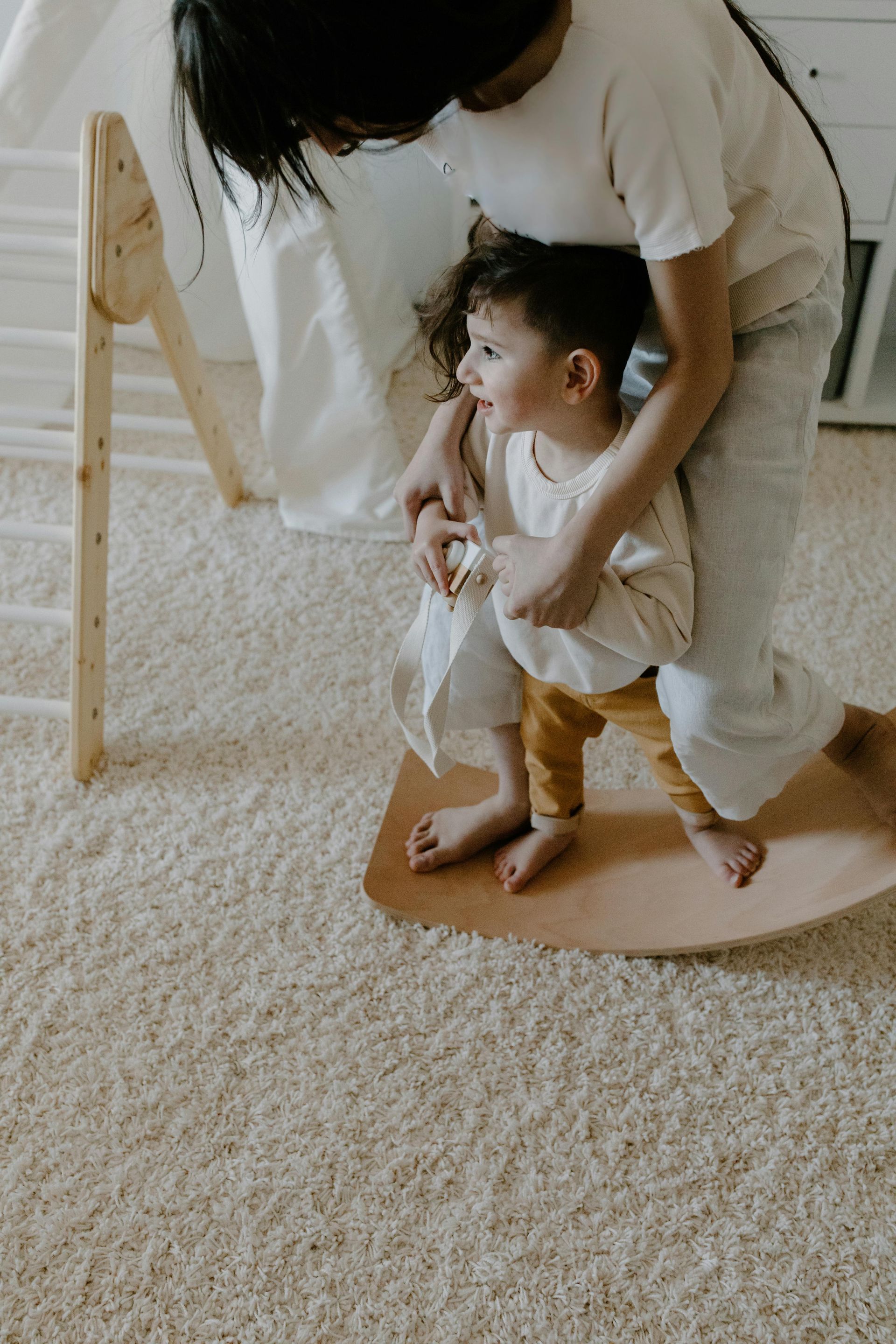 Person assists child on a wooden rocker in a light-colored, carpeted room, with a wooden climbing structure nearby.