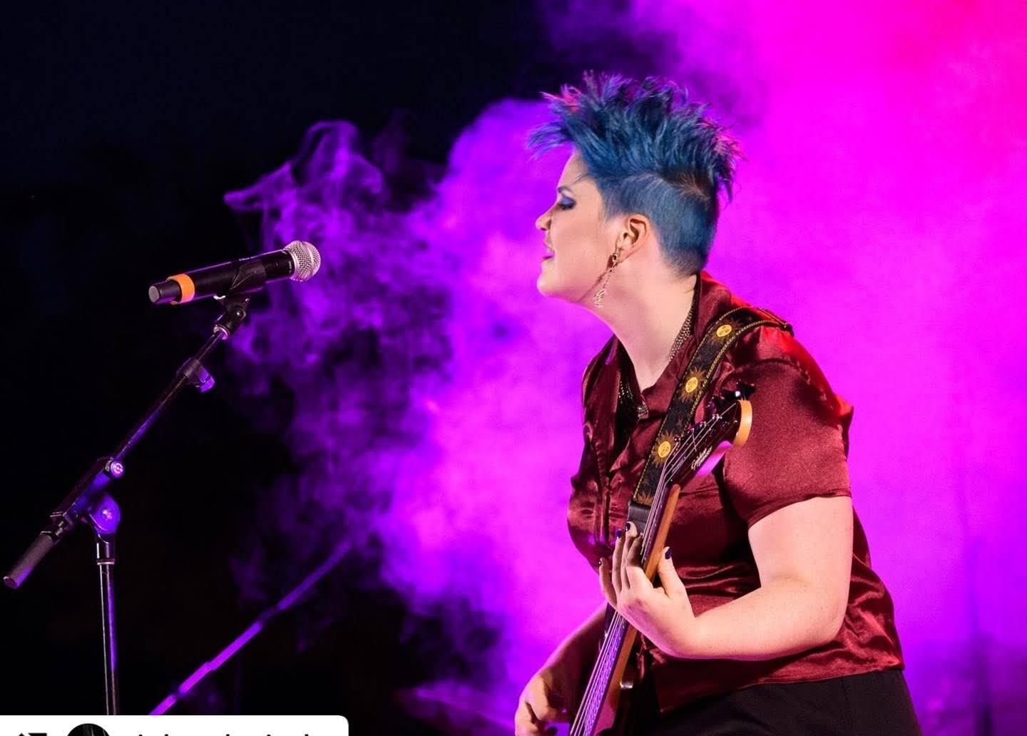 Woman with blue hair sings and plays guitar on stage, bathed in purple light.