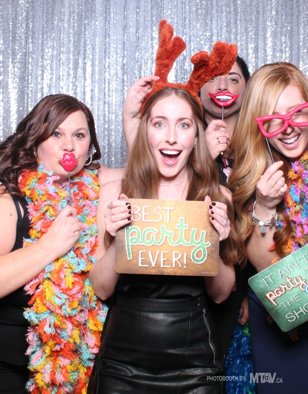 Four people posing with party props in front of a silver sequined backdrop; holding signs, lips and glasses.