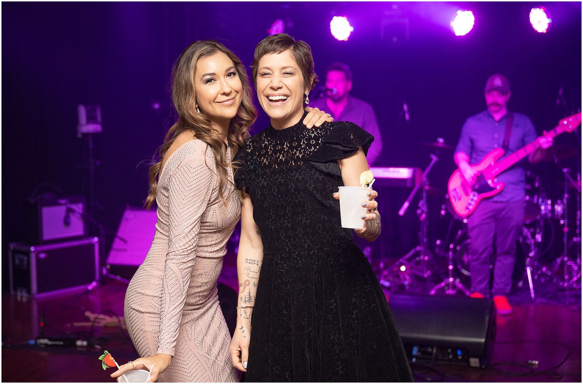 Two women smile at a party; band plays in the background, lit with purple lights.