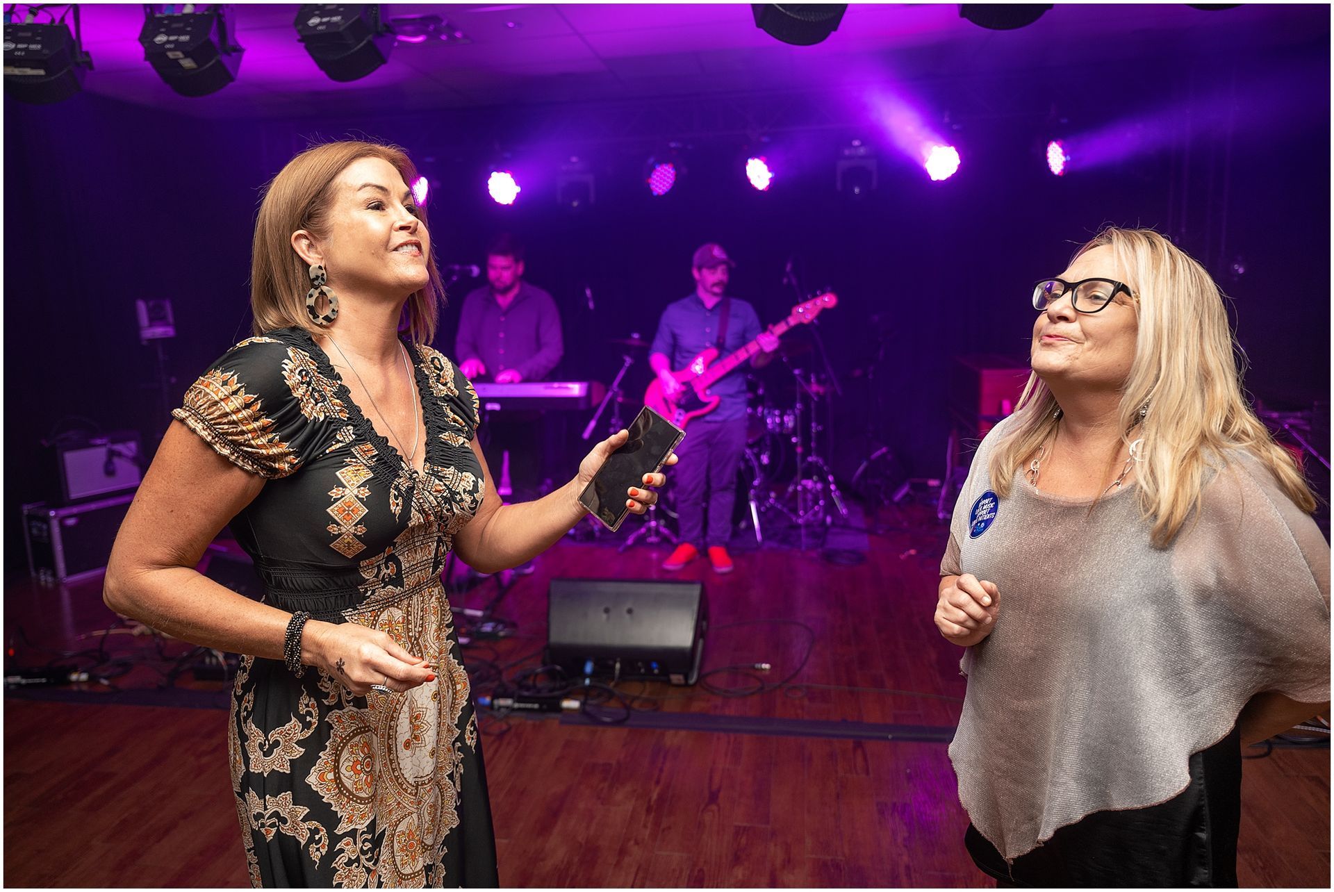 Two women on stage with a band playing; woman holding a phone, other smiling, both in front of a purple-lit stage.