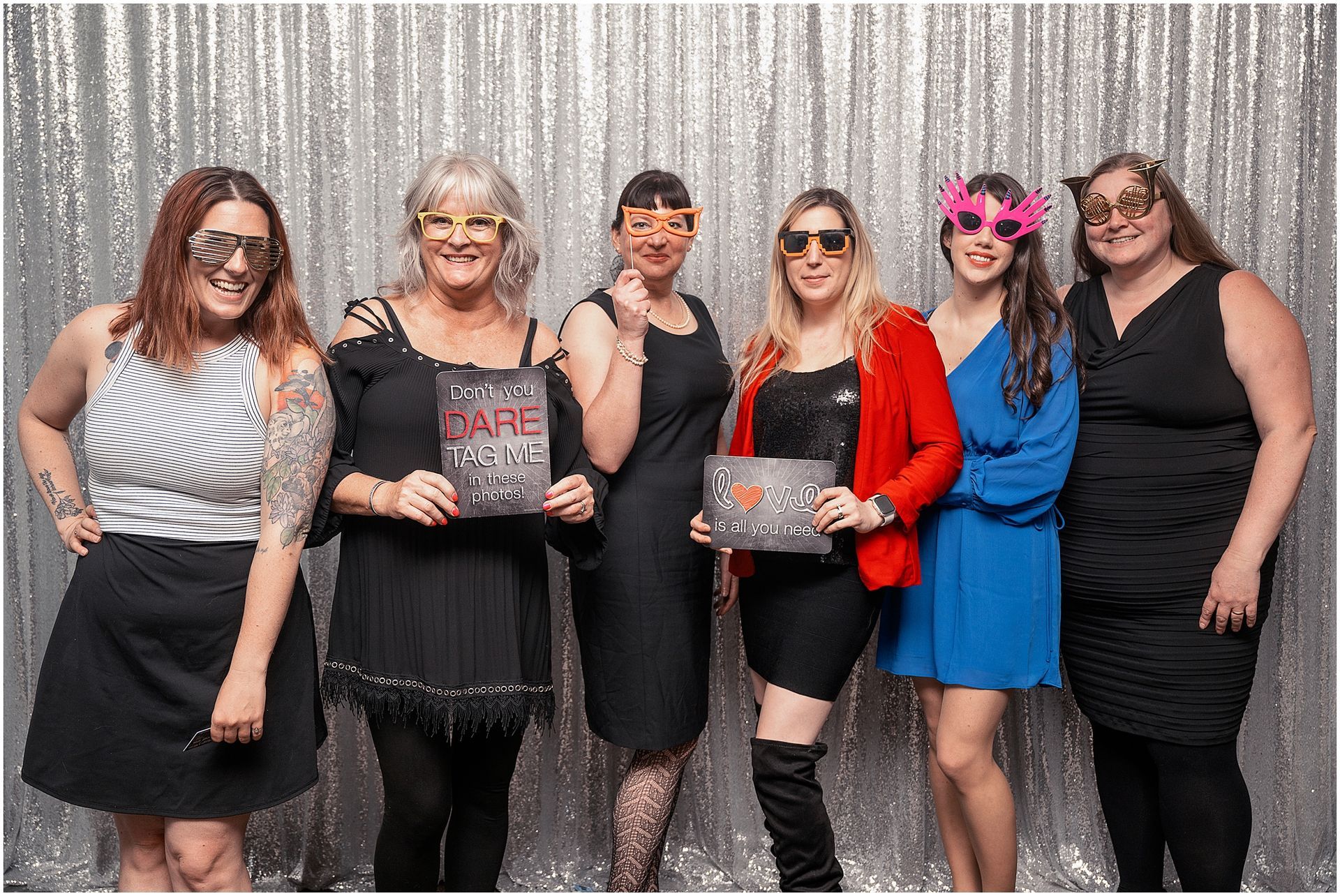 Six people pose in front of a silver backdrop, wearing costume glasses and holding signs, smiling.