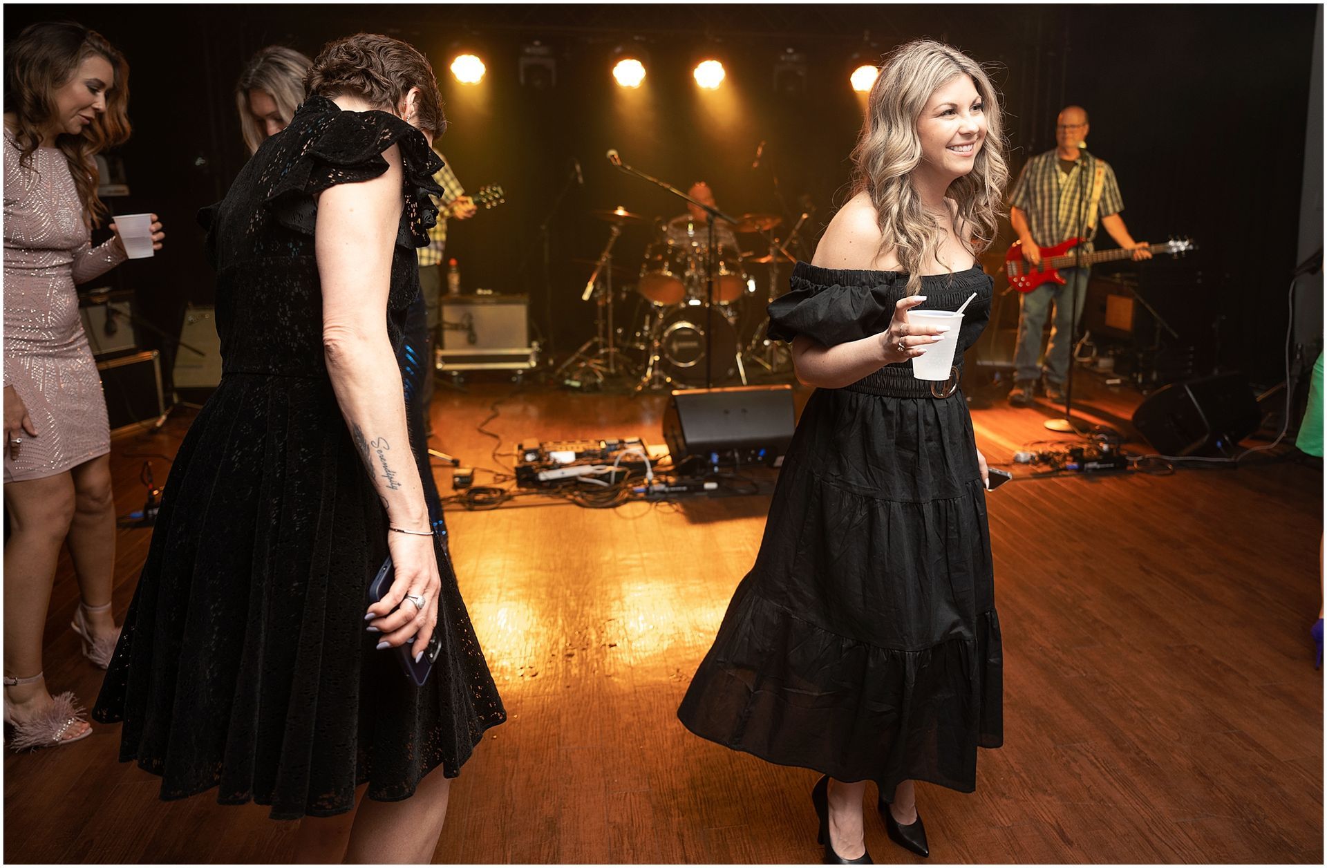 People dancing at a live music event. Woman in black dress holds cup, band plays on stage.