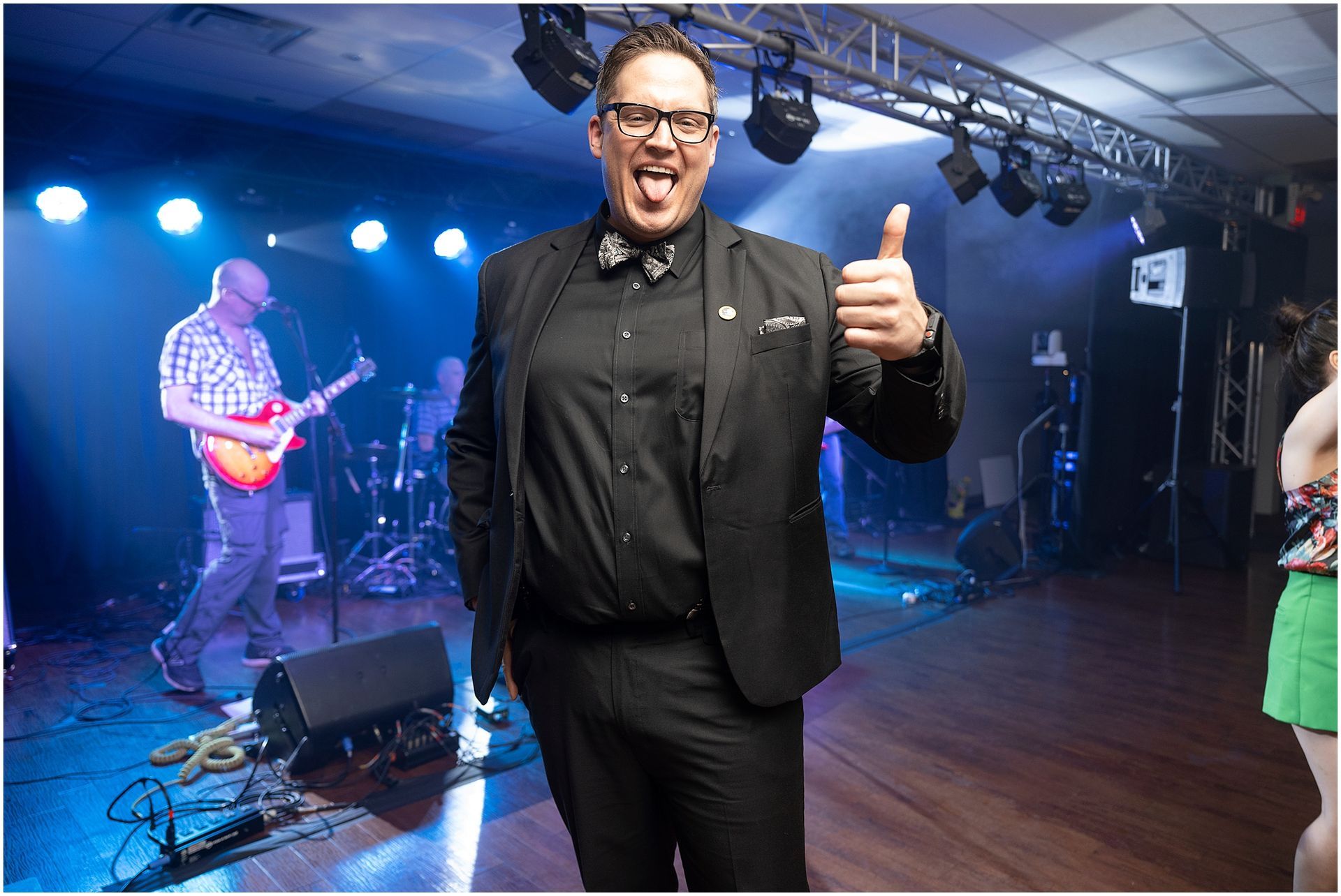Man in black suit giving thumbs-up on stage with band in background.
