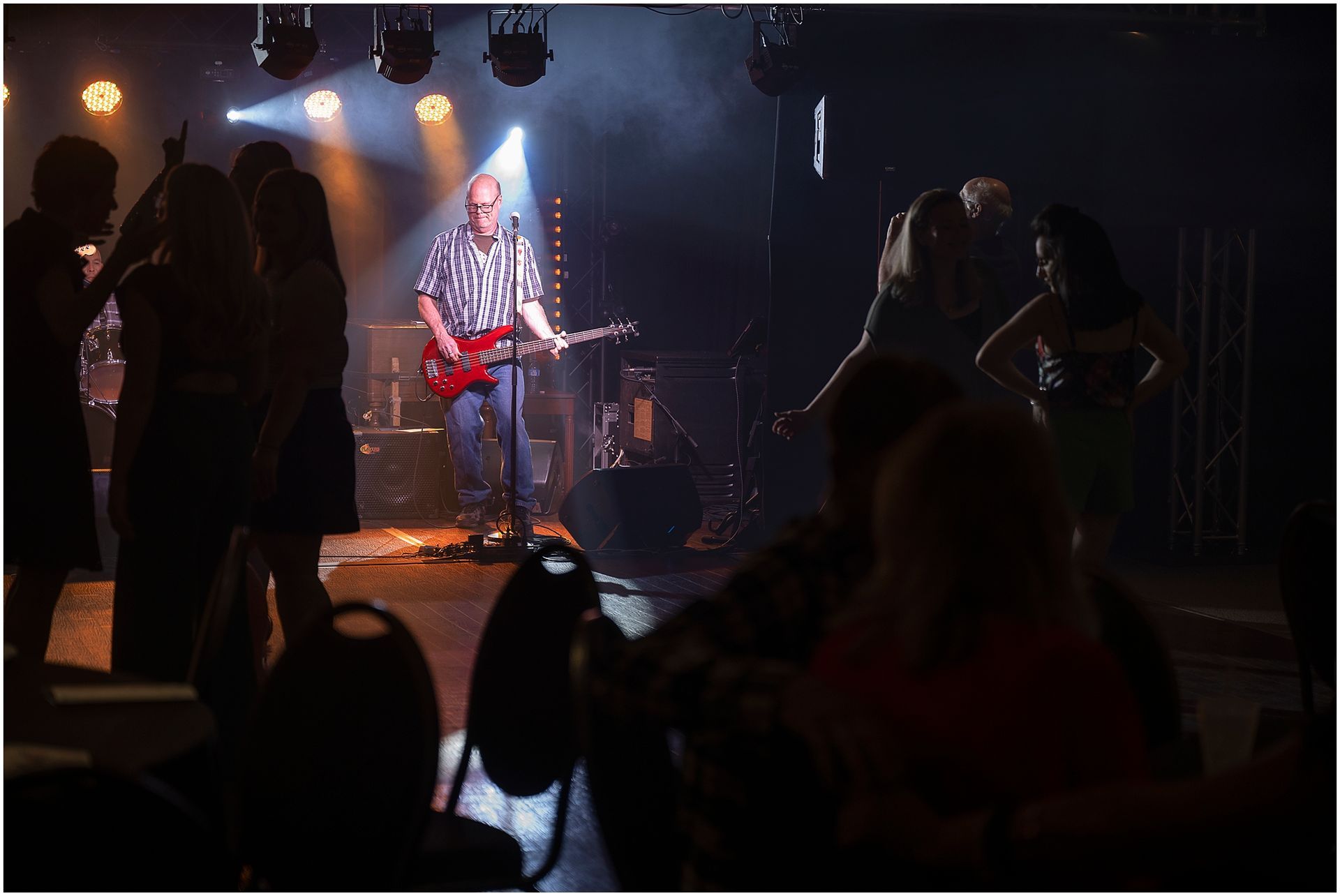 Band performing on stage in a dark club. People dance and watch. Stage lights shine on the band.