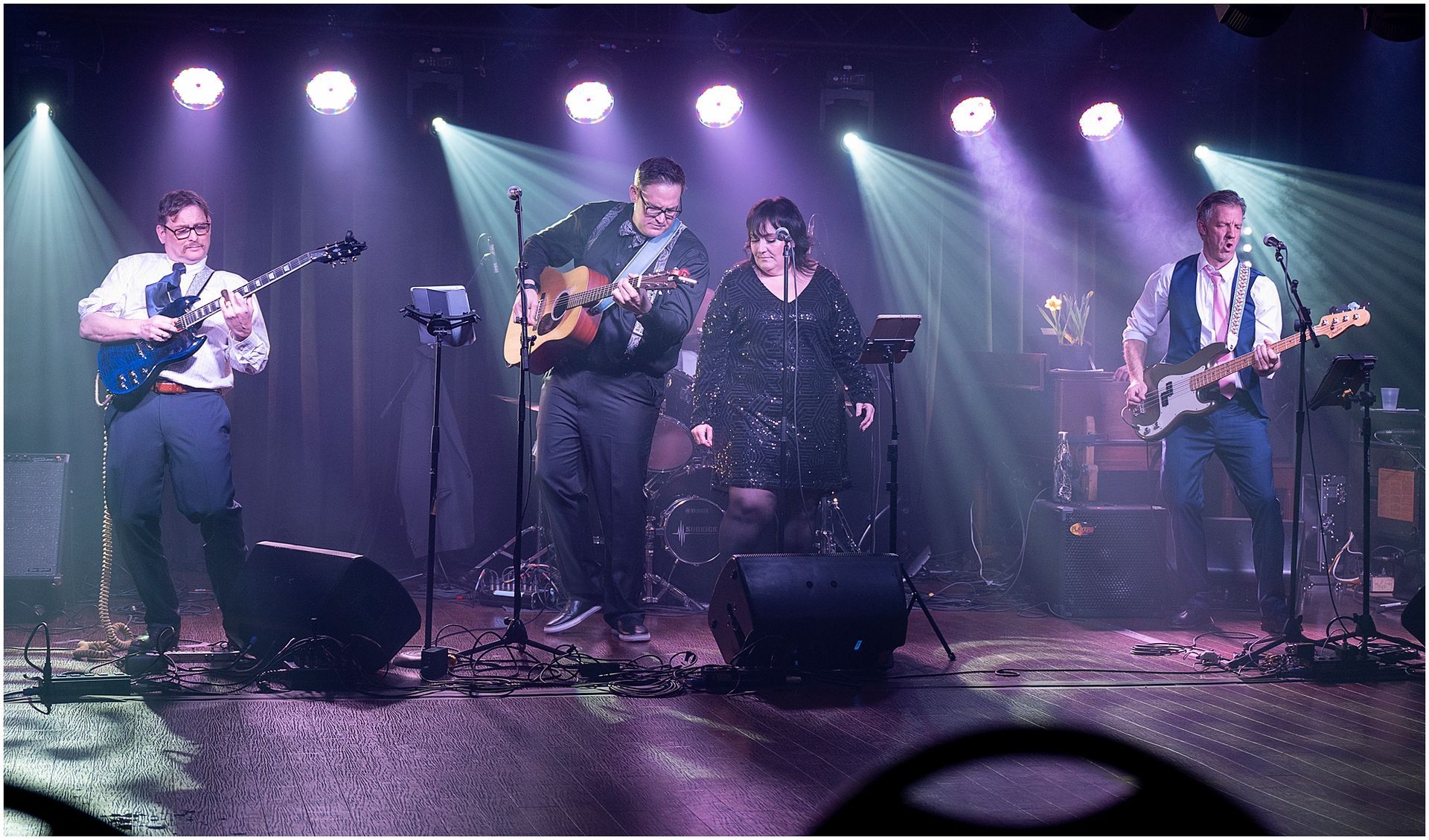 Band performing on stage, lit with purple lights. Instruments include guitars and keyboard. Singer in a sparkly dress.