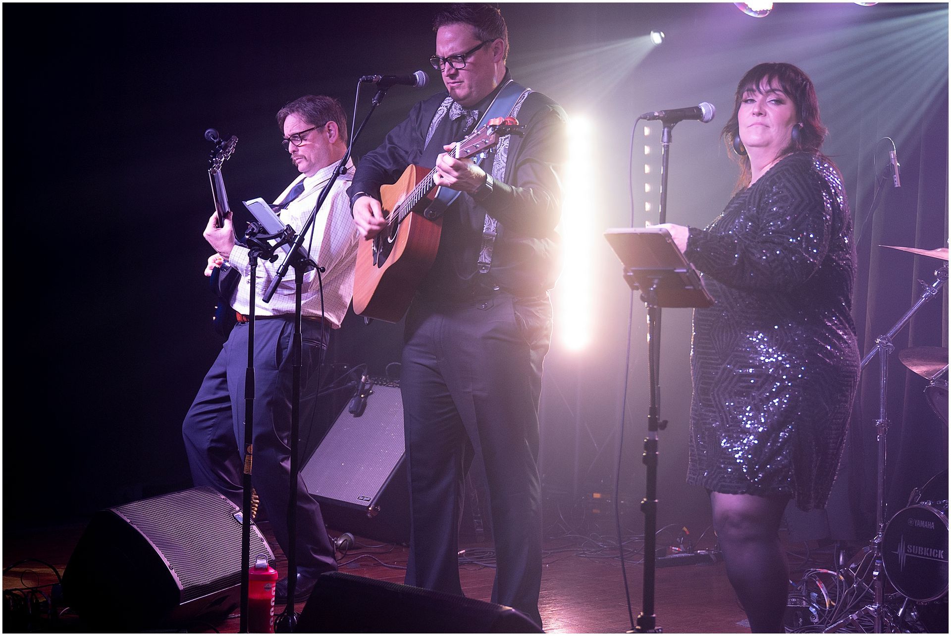 Band performing on stage under stage lights. Guitar, microphone, and a woman holding a tablet.