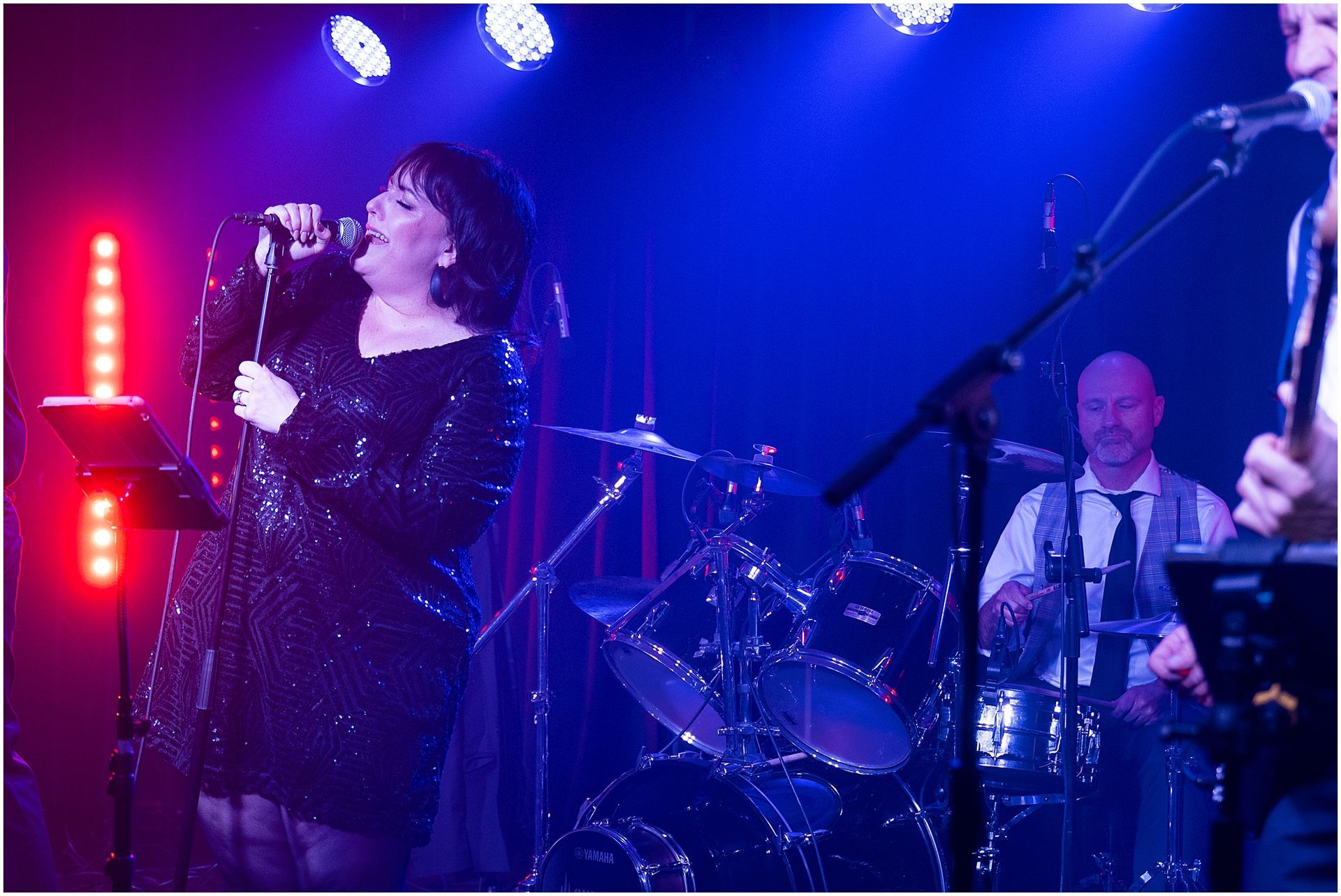 Woman singing with band on stage under blue and purple lights. Drummer visible.