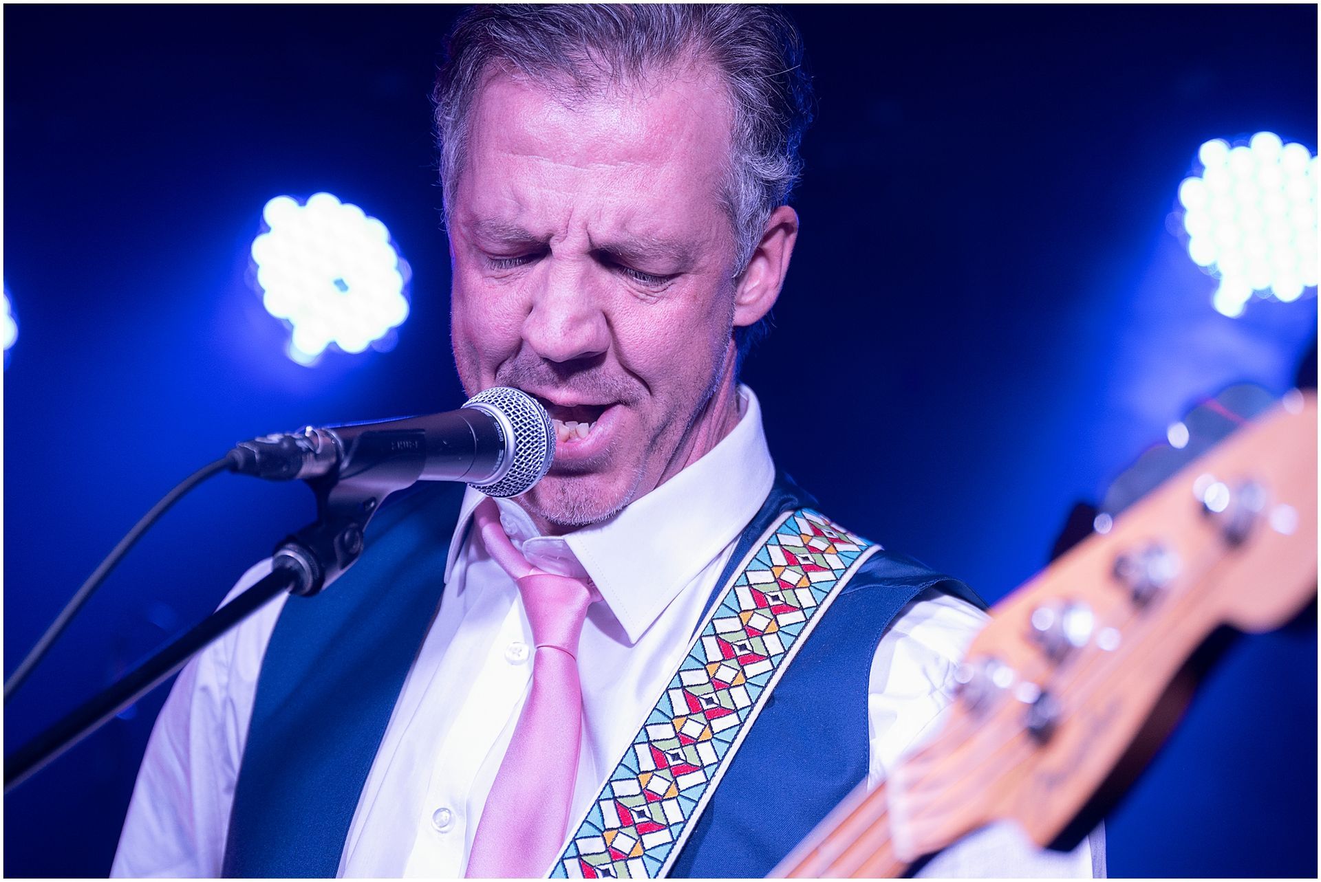 Man singing into a microphone, playing bass guitar on stage, wearing a vest and pink tie.