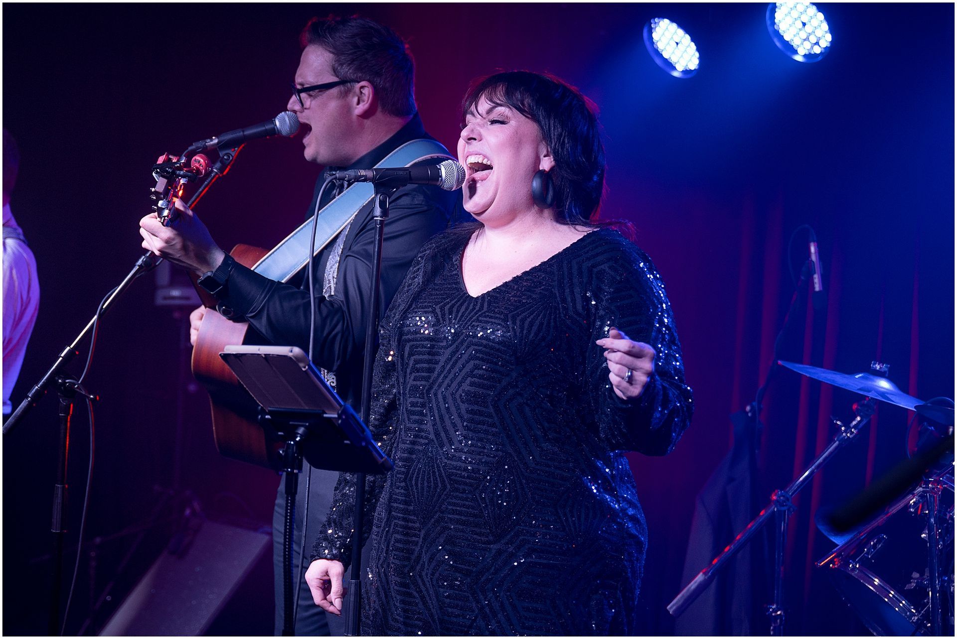 Man and woman singing into microphones on stage, man plays guitar, woman wears a black sequined dress.