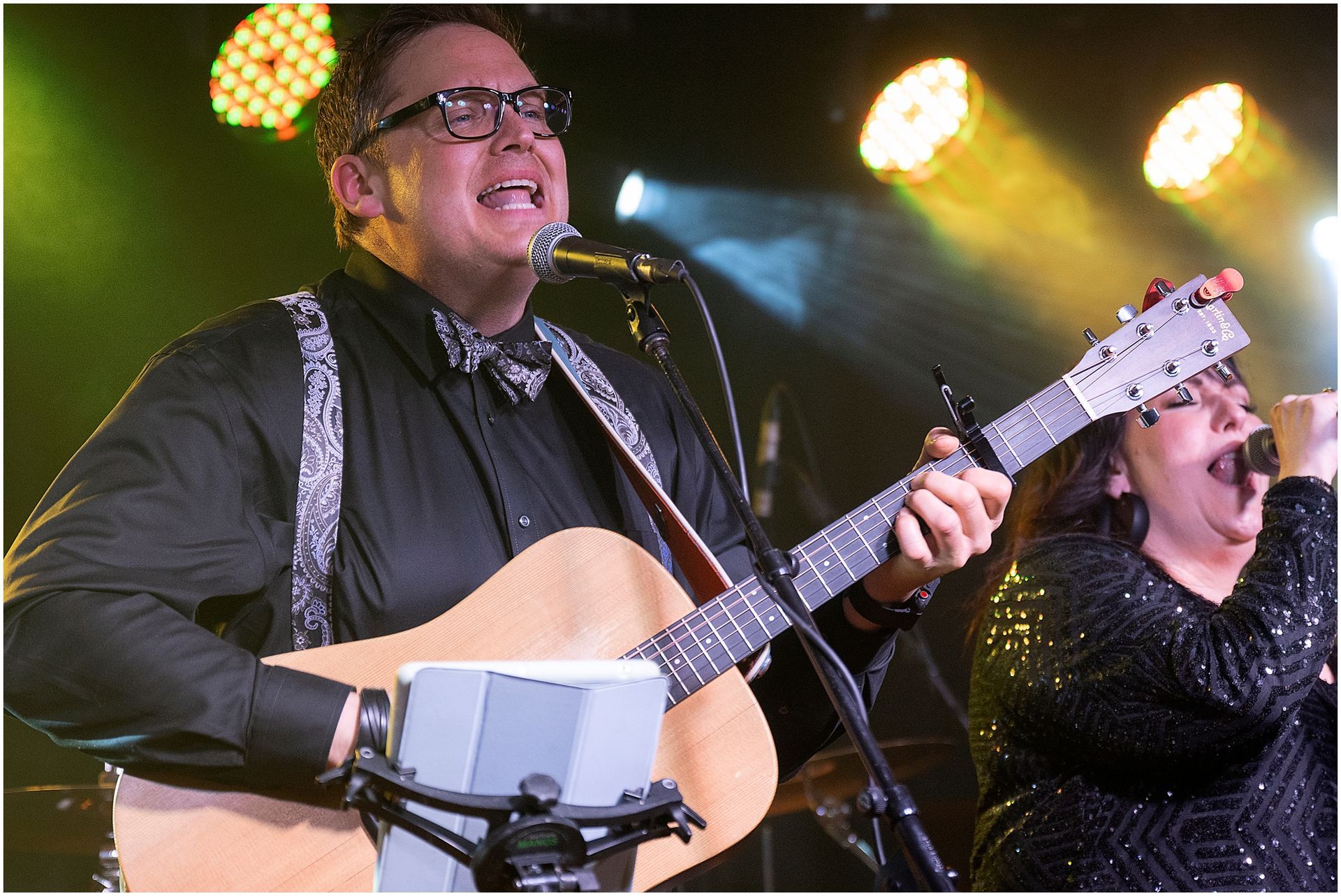 A man playing guitar and singing, joined by a woman singing, on a stage with stage lighting.