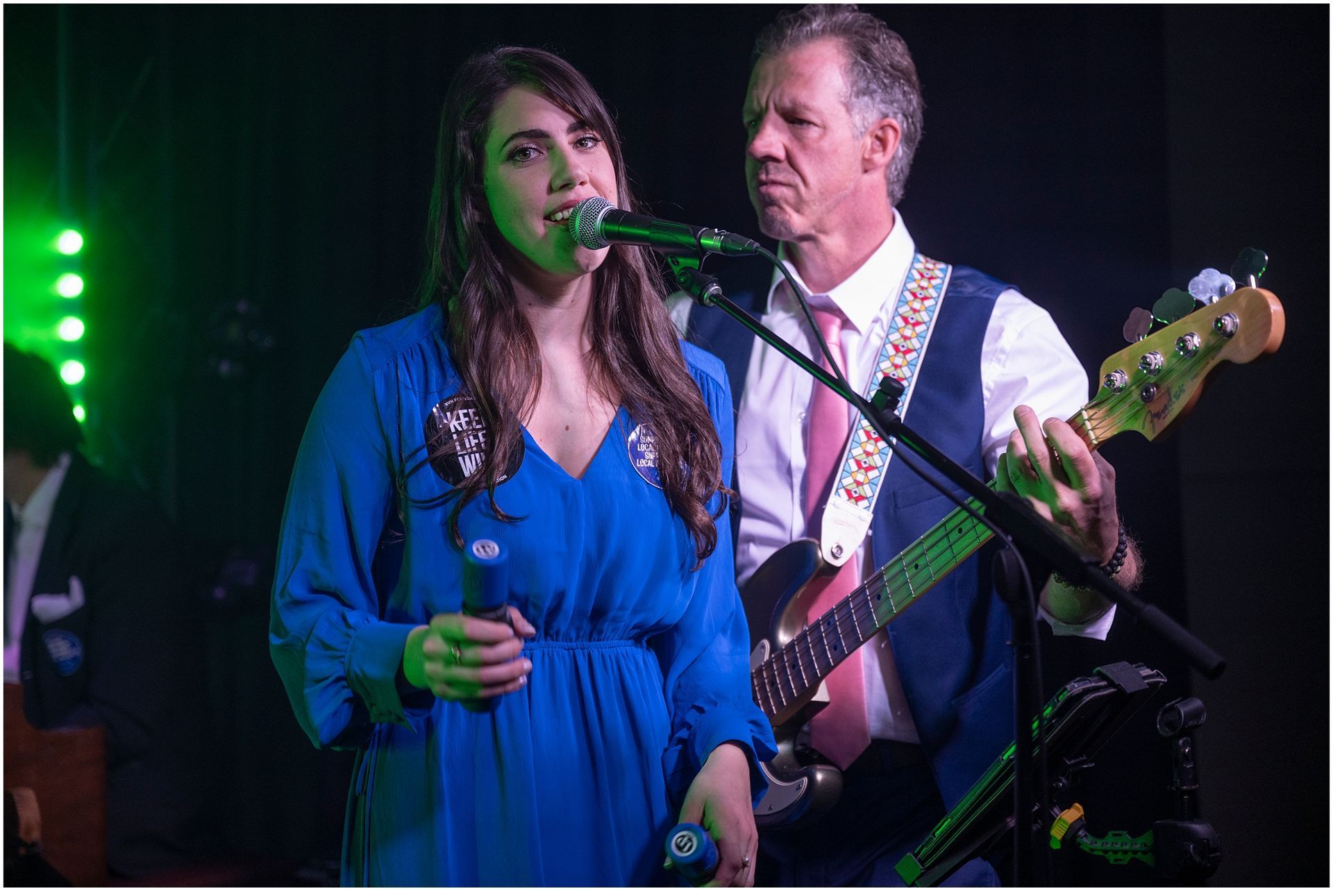 Woman in blue dress singing into a microphone, man playing bass guitar behind her on stage.
