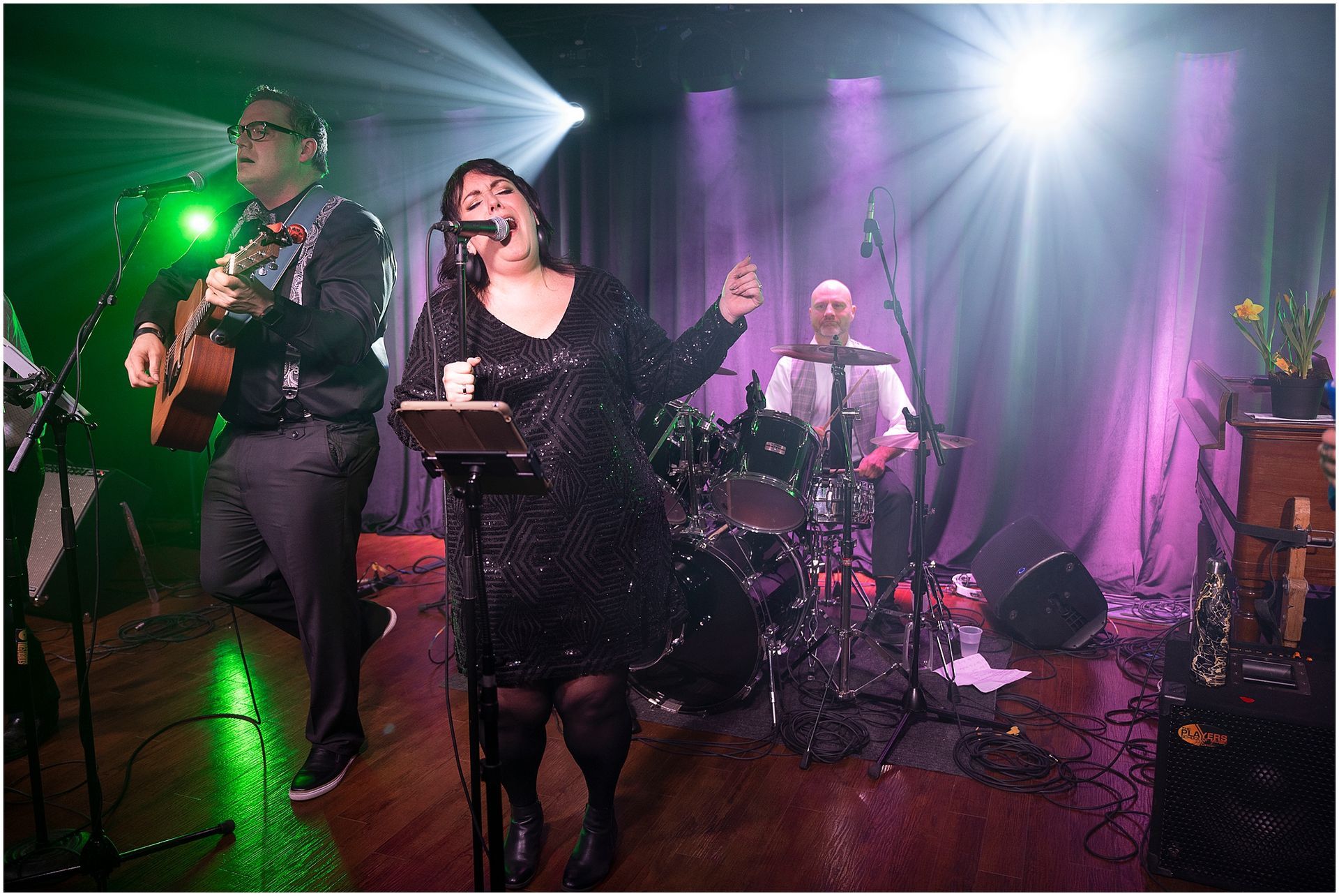 Band performing live on stage; woman singing with microphone, man playing guitar, drummer in background.
