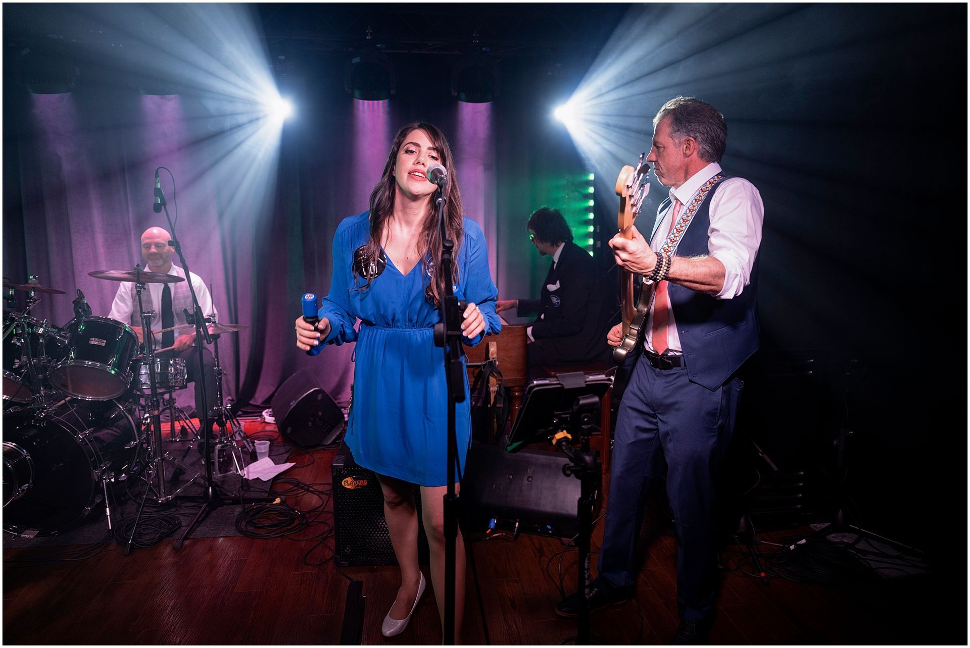 Band performing on stage, singer in blue dress, bassist, drummer, keyboardist, spotlight.