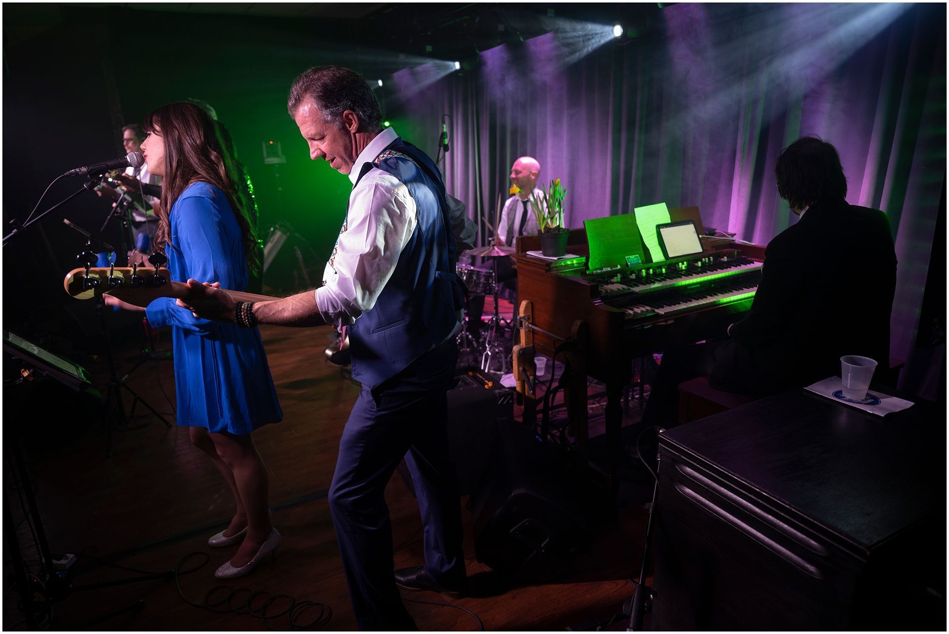 Band performing on stage with colorful lighting. Woman sings and plays guitar, man plays guitar, others on keyboard and drums.