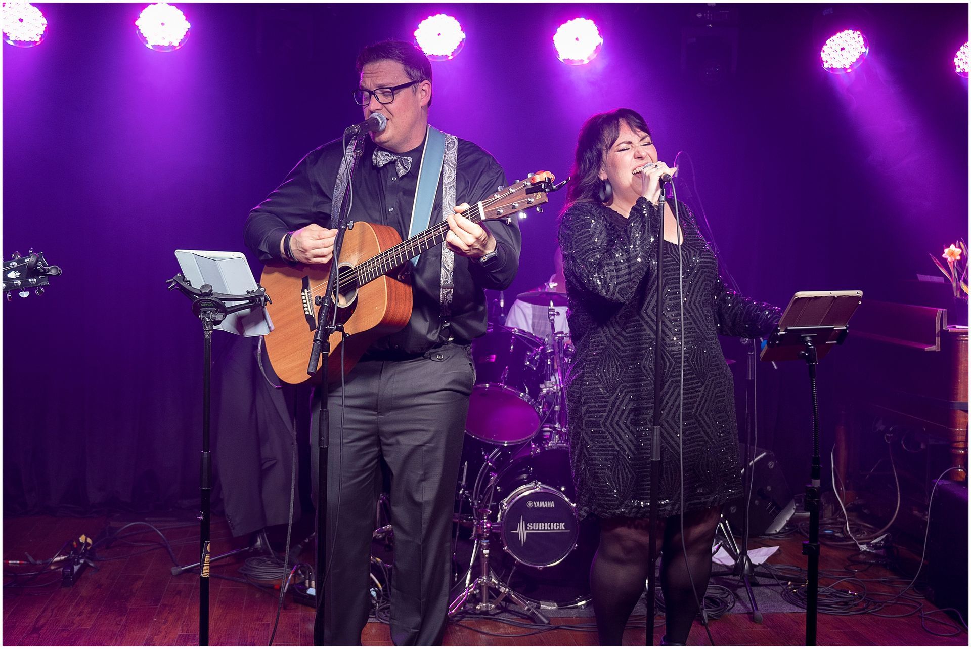 Two musicians on stage: man playing guitar, woman singing. Purple stage lighting.