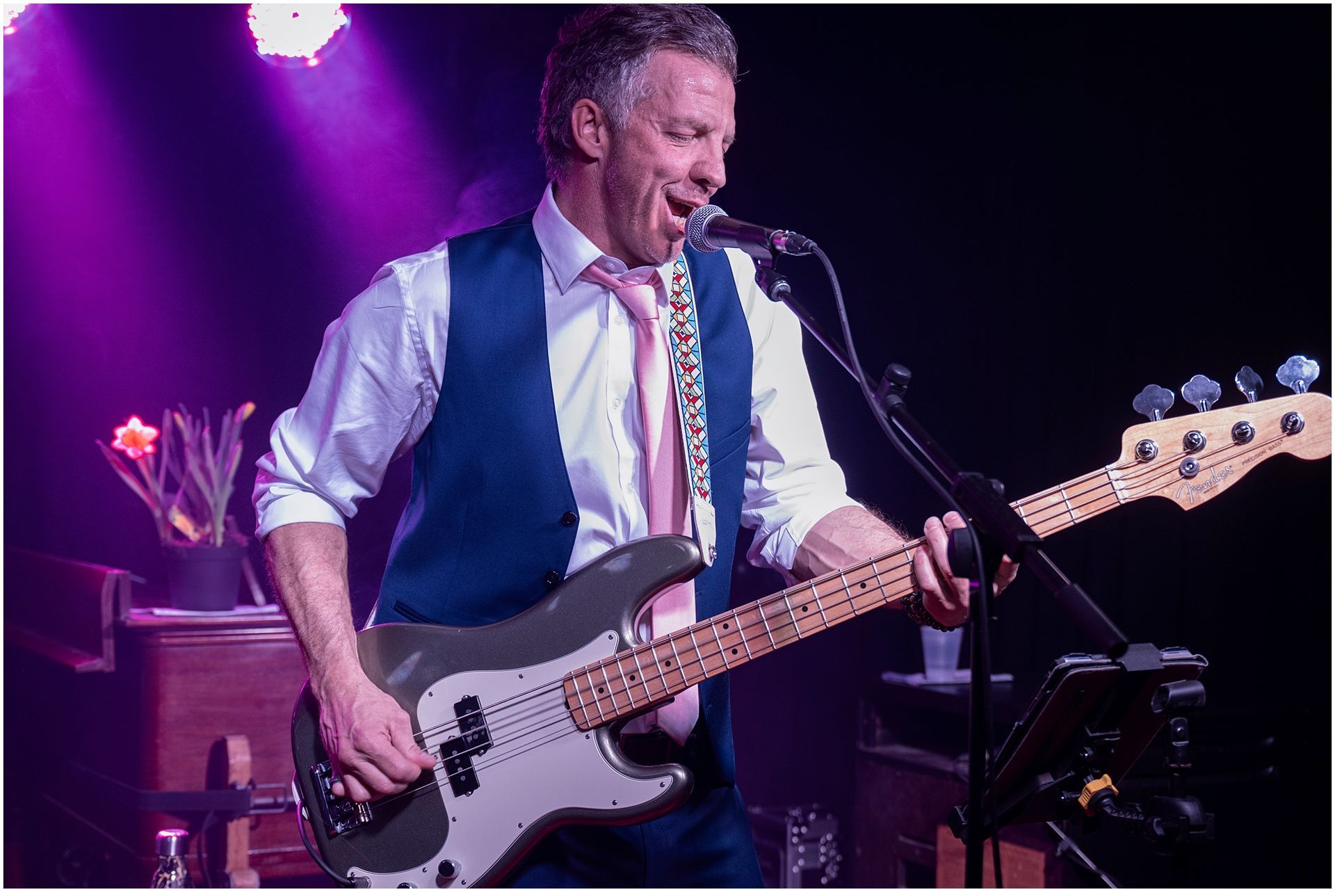 Man playing bass guitar and singing into a microphone on stage, under purple lighting.