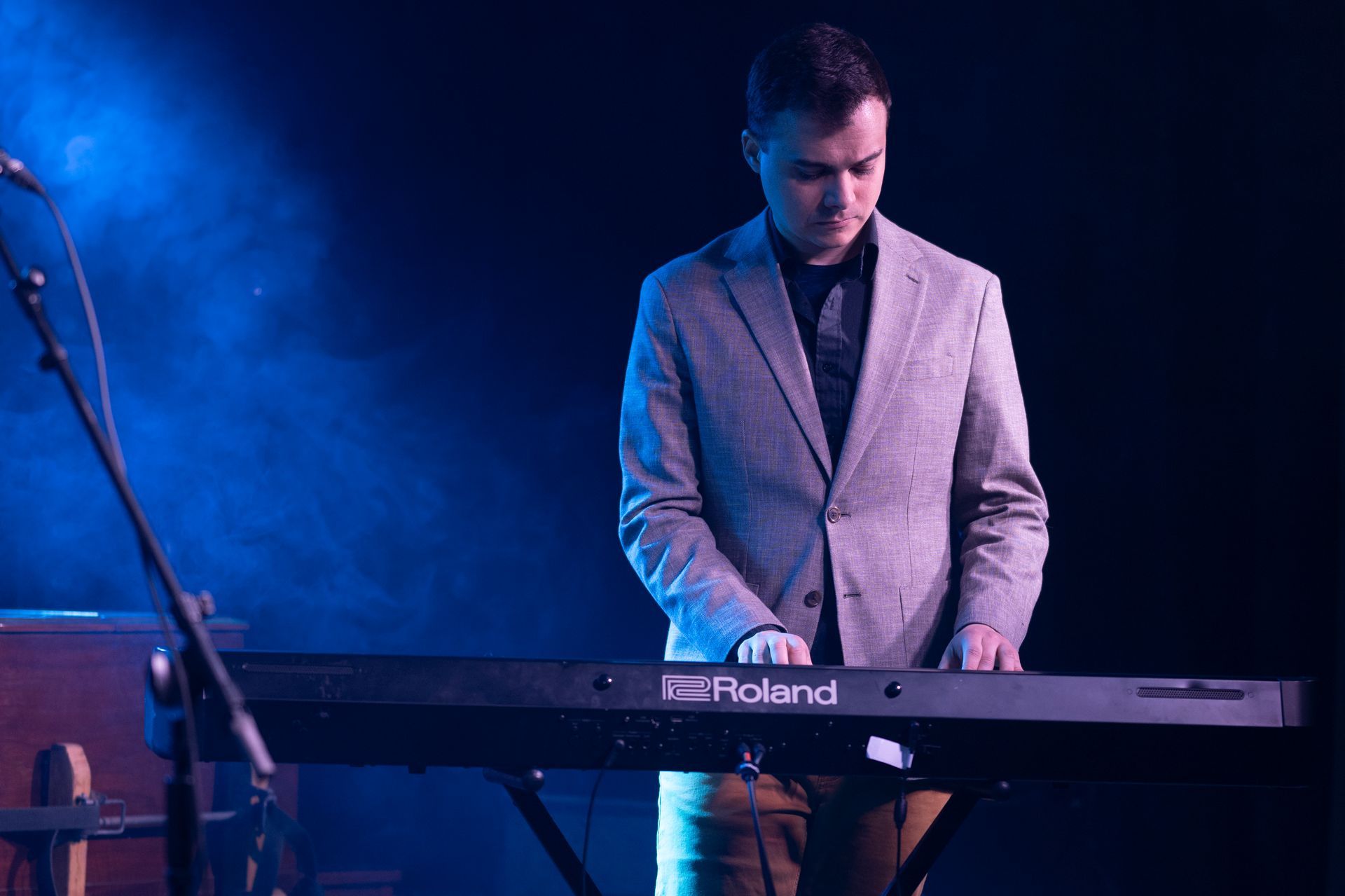 Person in a blazer playing a Roland keyboard on stage, illuminated by blue light.
