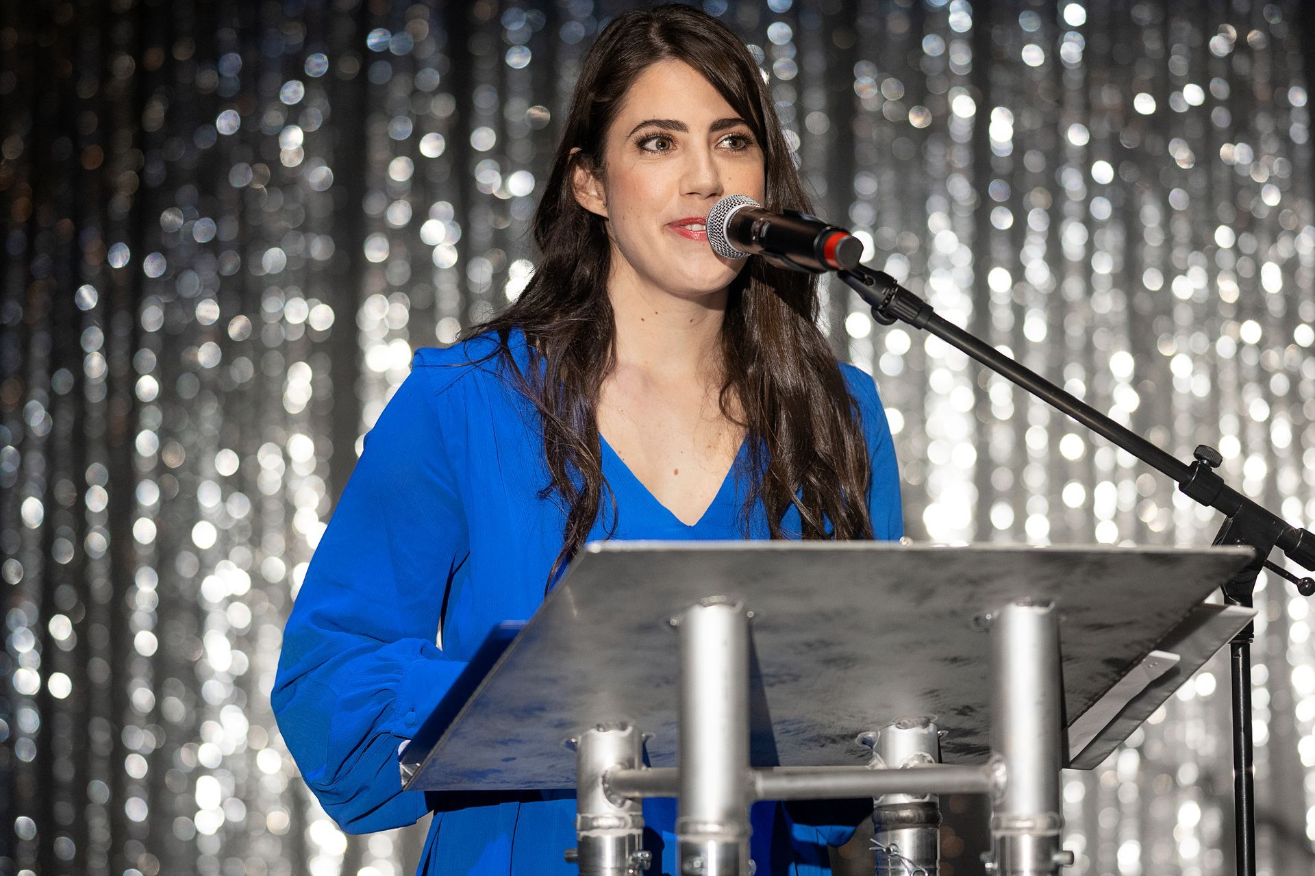 Woman in a blue dress speaks at a podium with a microphone. Silver background.