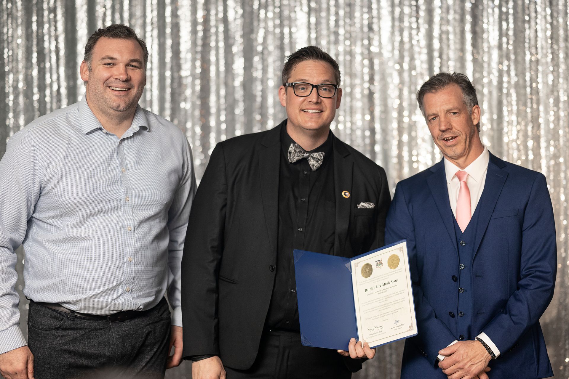 Three men stand in front of a silver backdrop. One holds a certificate, others smile.