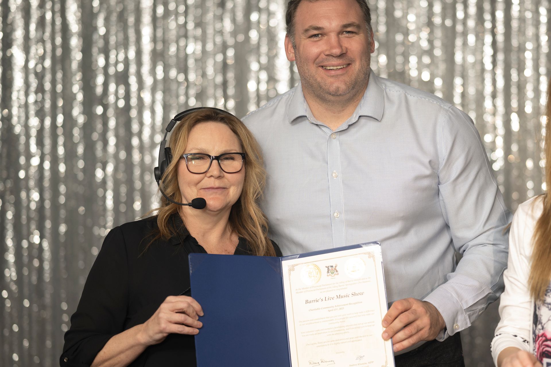 Two people holding a certificate, woman with headset smiling, silver backdrop.