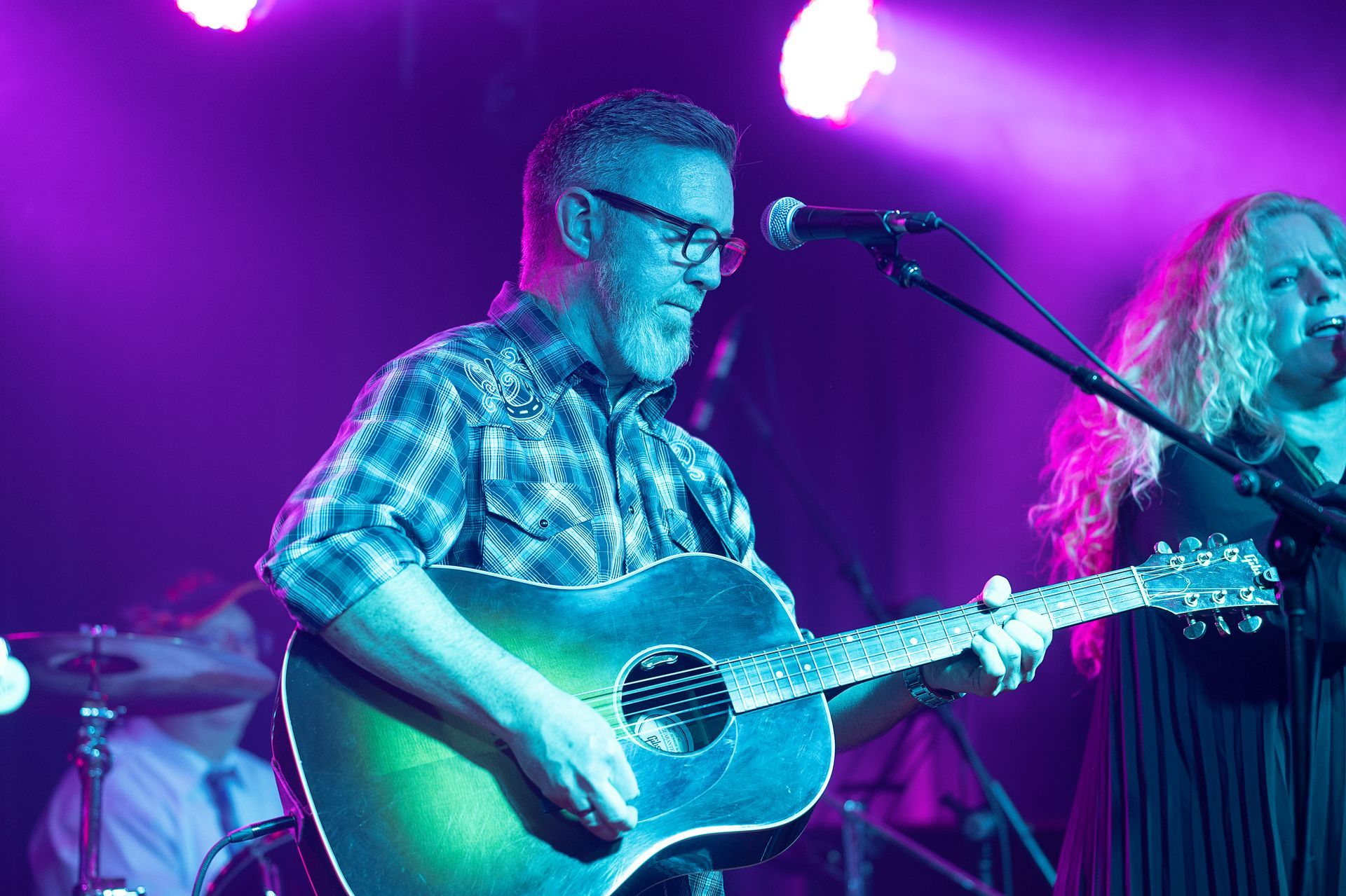 Man with glasses plays guitar on stage, singing. Purple and blue stage lighting.