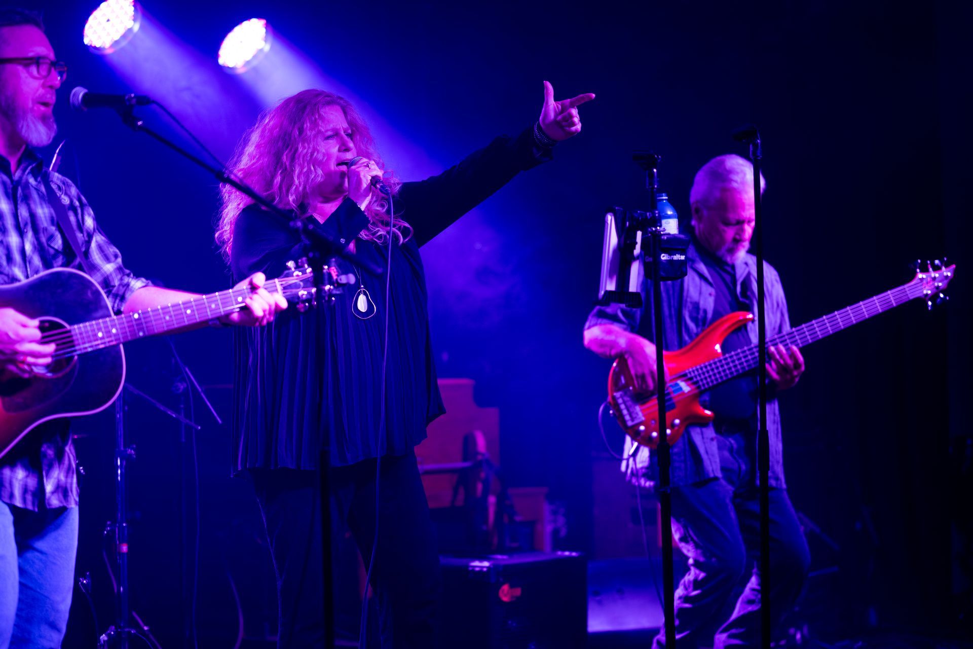 A band performing on stage under blue lights. Singer gestures, guitarist to the left, bassist on the right.