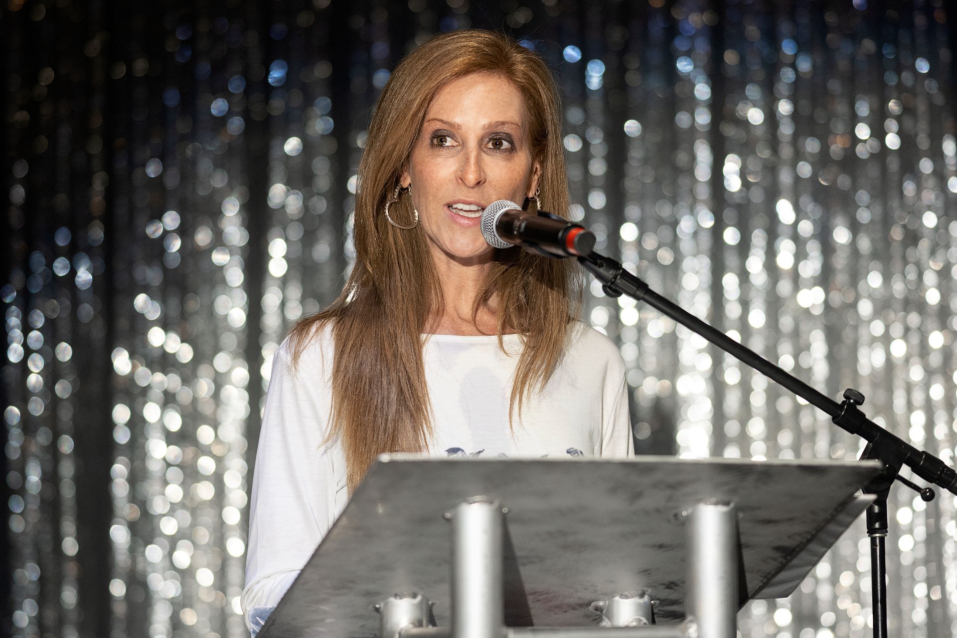 Woman speaking at a podium with microphone, in front of a shimmery silver background.