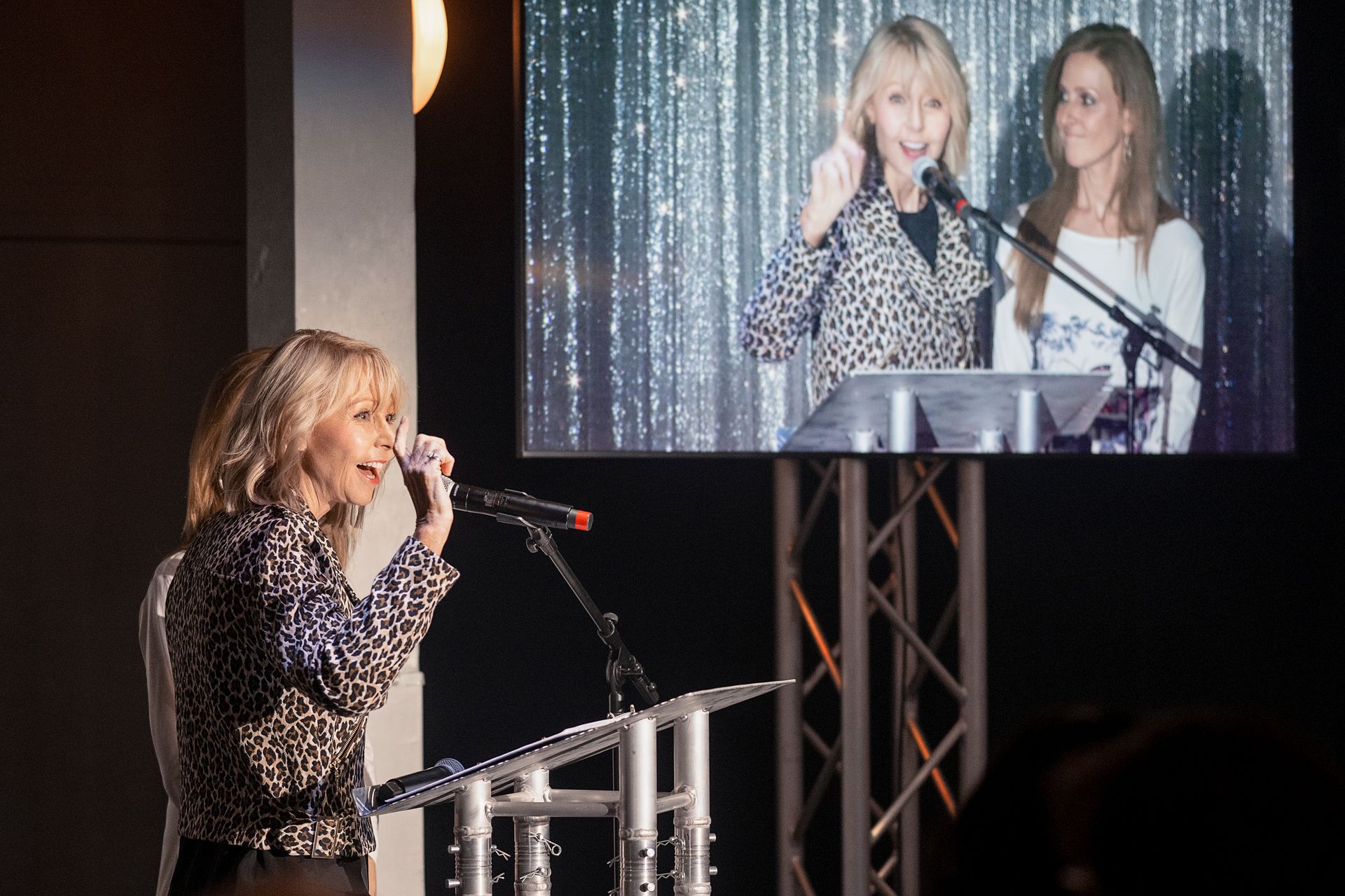 Woman speaking at podium, video screen shows her and another woman on stage.