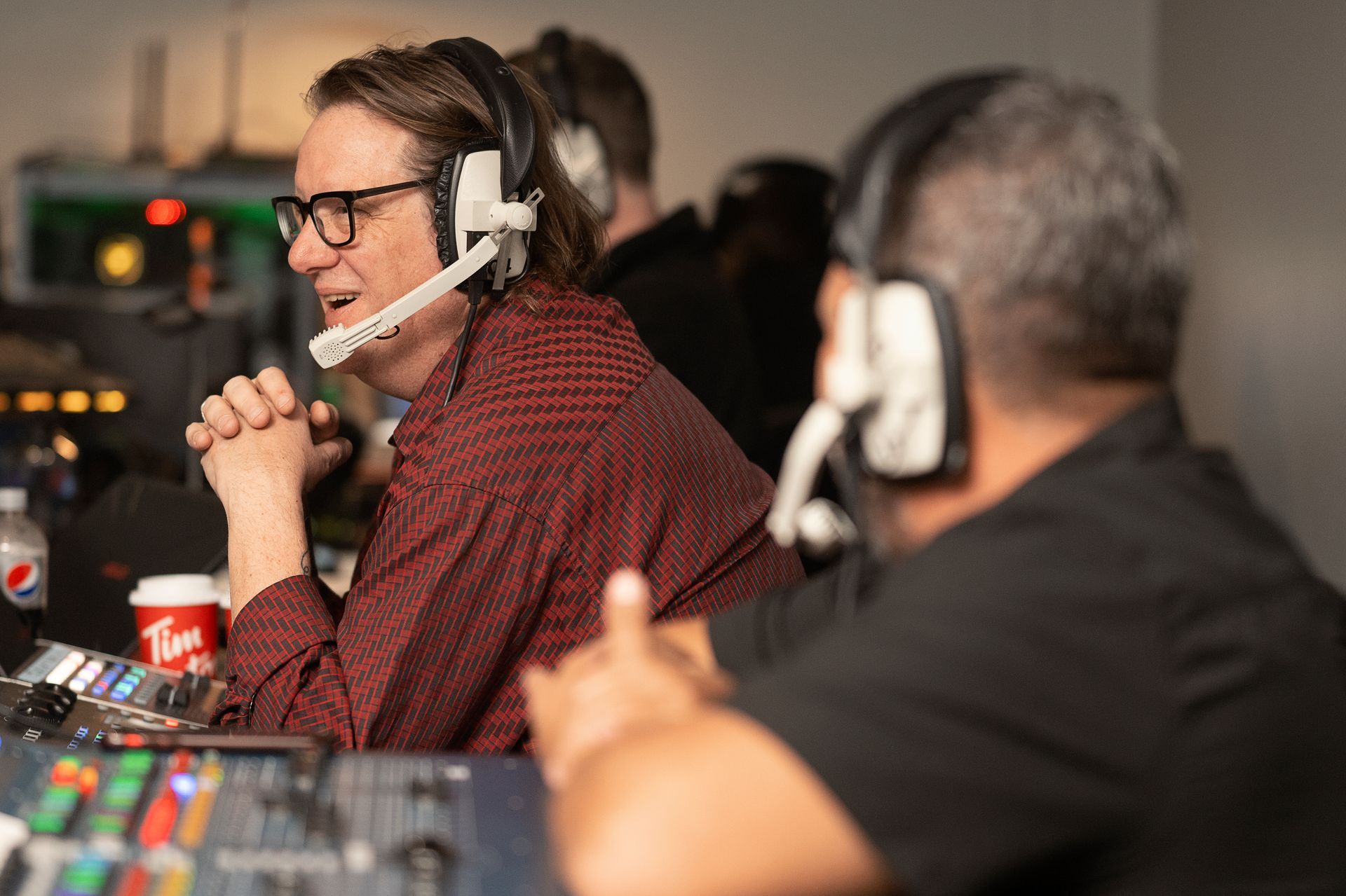 Man with glasses and headset at a control panel, looking toward another man in a headset.